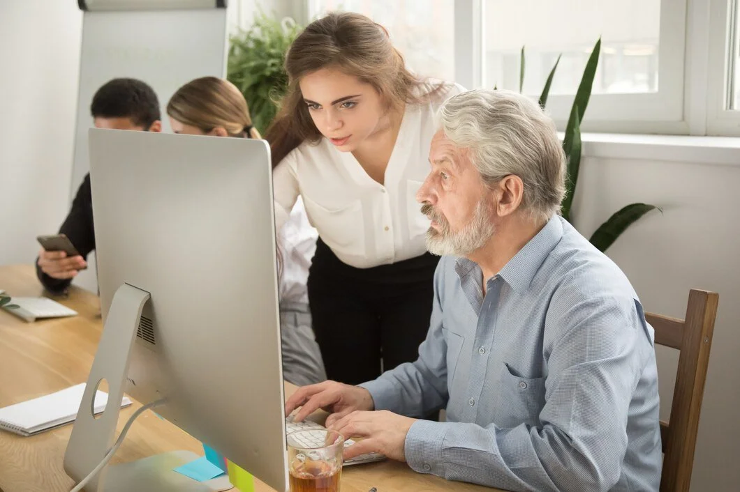 A young woman is explaining something on a computer to an older man in an office setting. The man is focused on the screen while the woman points at it. Other colleagues are seen in the background, one looking at a smartphone. The office has plants and stationery on the desk.