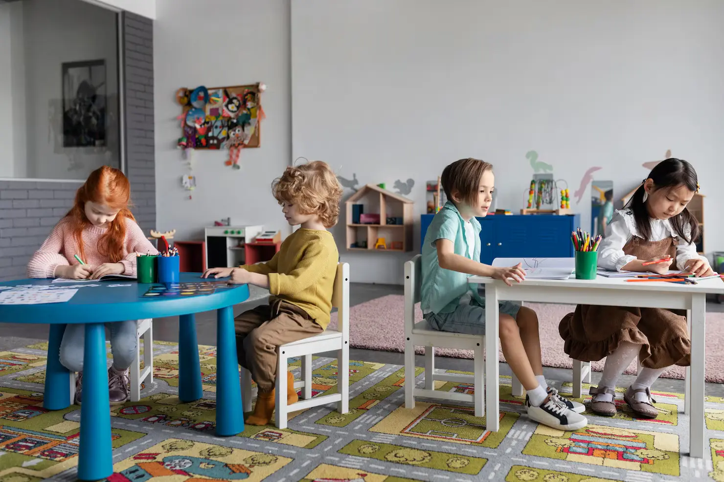 Four young children are seated at tables in a kindergarten classroom, focused on drawing and coloring. A girl with red hair is coloring at a blue table, while the others sit at white chairs, working with crayons and papers. The room has colorful decorations and a play area in the background.