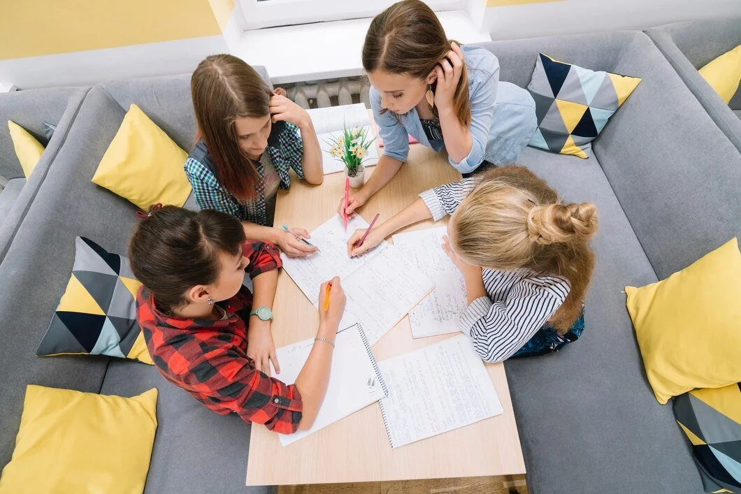 Four girls are working together on their homework around a table. They are focused on writing and discussing, with notebooks and pens in front of them. The room is cozy, with yellow and gray cushions on the couch, and a small plant in the center of the table.