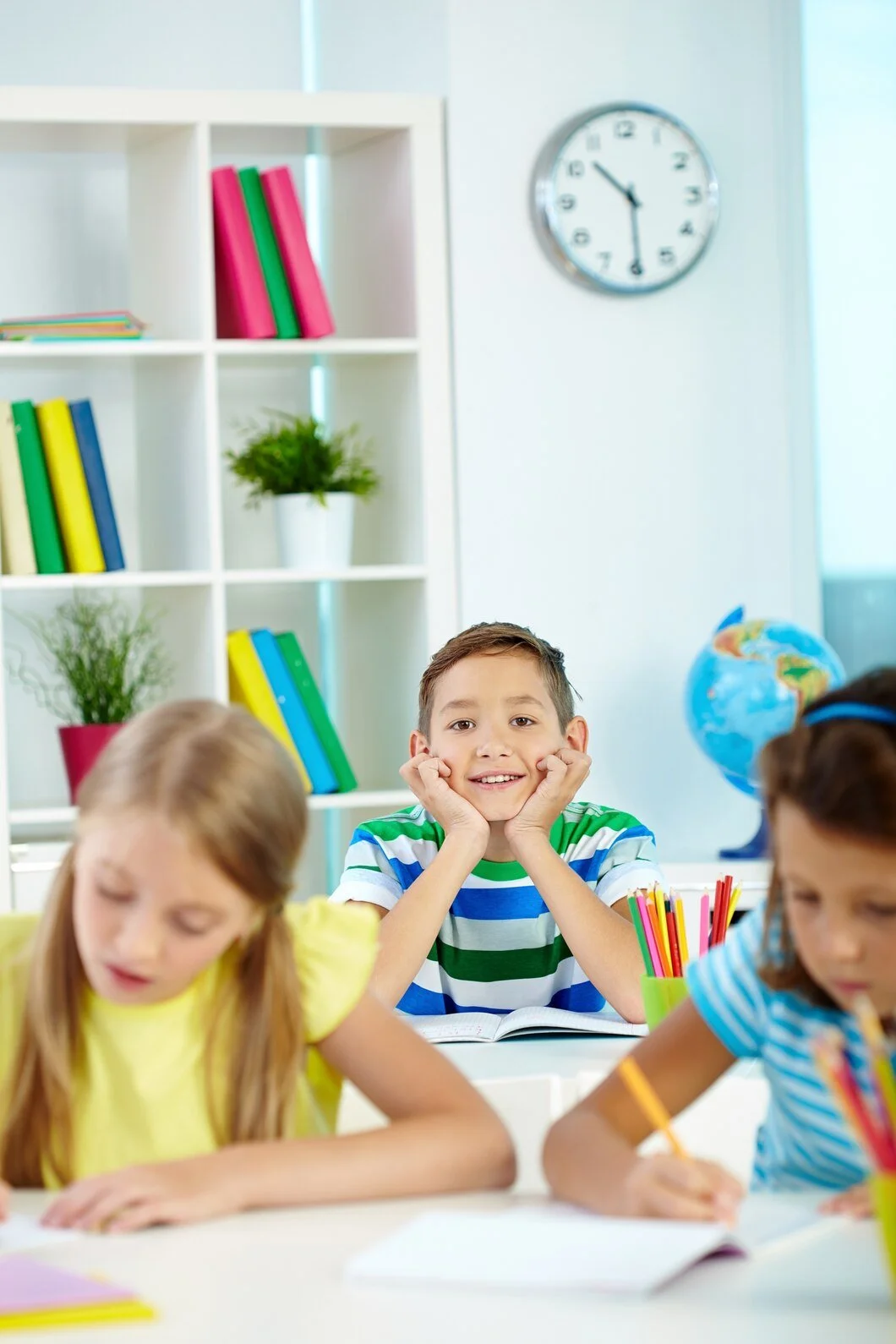 A young boy smiling with his chin resting on his hands in a classroom, surrounded by classmates writing in their notebooks. Books are stacked on a shelf, and a globe is visible in the background.