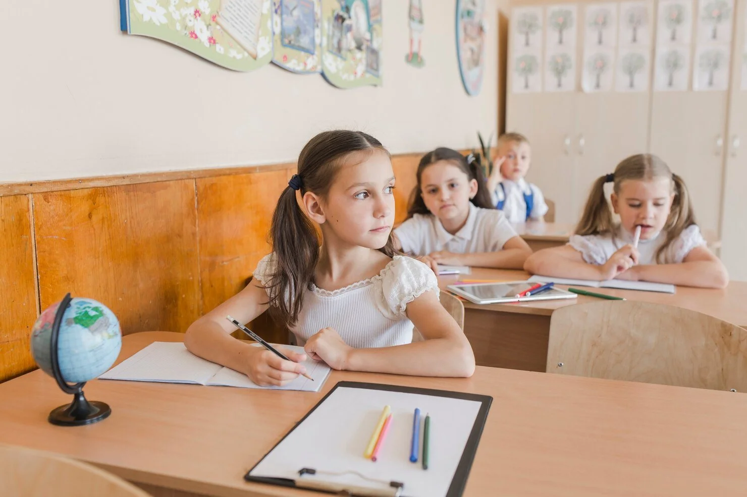 A young schoolgirl sitting at a desk, holding a pencil and looking away thoughtfully. Other children can be seen in the background, working quietly at their desks. A globe and colorful crayons are visible on the table.